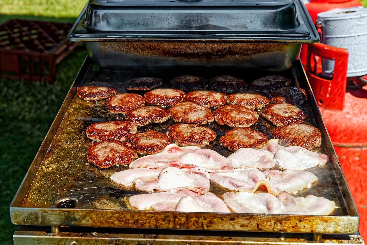 Burger patties and rashers of bacon cooking across an outdoor flat-top griddle in daylight